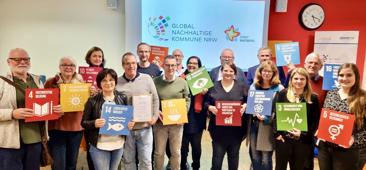 16 Frauen und Männer sehen nebeneinander vor einem Logo der Global Nachhaltigen Kommunen und halten farbige Schilder, auf denen die globalen Nachhaltigkeitsziele aufgeführt sind.