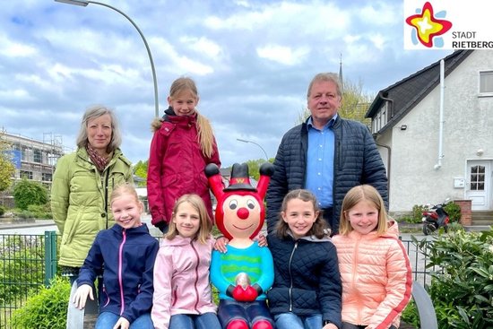 Lia, Jule, Ida, Pia und Marlene sitzen unter freiem Himmel auf einer Bank - die Rieti-Figur »Westi« in der Mitte. Kunstwerkstatt-Leiterin Karin Wolf und Wilfried Dörhoff von der Stadt Rietberg stehen links und rechts daneben und blicken ebenfalls in die Kamera.