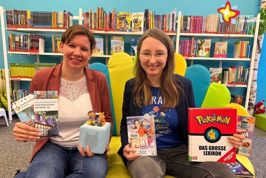 Julia Bröckling und Jennifer Bader sitzen in der Kinderecke auf einem Sofa und zeigen einige der beliebtesten Medien. Im Hintergrund ist ein großes Bücherregal zu sehen.