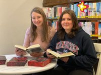 Zwei Frauen sitzen mit jeweils einem Buch in den Händen hinter einem kleinen runden Tisch und vor einem Regal voller bunter Bücher.