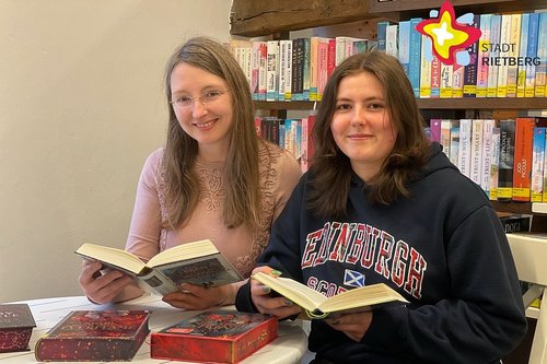 Zwei Frauen sitzen mit jeweils einem Buch in den Händen hinter einem kleinen runden Tisch und vor einem Regal voller bunter Bücher.