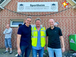 Bürgermeister Andreas Sunder, Thomas »Tobi« Kofort und Thomas Trendelbernd stehen nebeneinander unter dem Schild mit der Aufschrift "Sportheim der Druffeler Sportgemeinschaft"