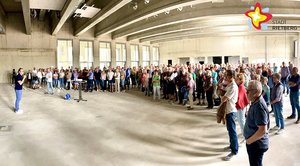 In einer großewn Halle mit Betonwänden stehen hunderte Menschen im Halbkreis um Bürgermeister Andreas Sunder, der - links im Bild - zu ihnen spricht.