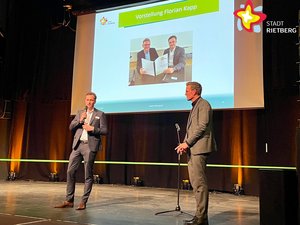 Florian Kapp steht mit einem Mikrofon in der Hand auf der Bühne, rechts im Bild Bürgermeister Andreas Sunder. Im Hintergrund wird ein Foto der zwei auf eine große Leinwand projiziert.