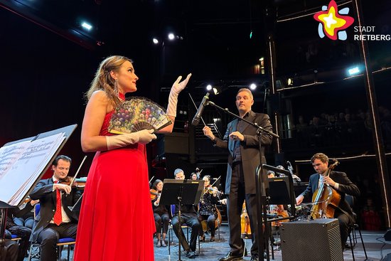 Sopranistin Eva Hartová steht in einem langen roten Kleid auf der Bühne und singt. Sie hält einen Fächer in der Hand. Neben ihr steht Dirigent Johannes Klumpp.