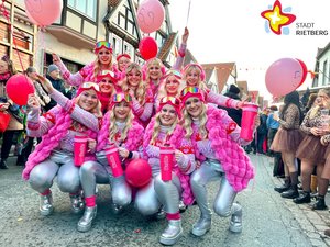 Eine Gruppe Frauen in silbernen Hosen und pinkfarbenen Westen lachen unter blonden Perücken und Skibrillen auf der Stirn in die Kamera.