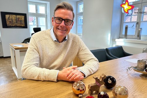 Der Bürgermeister sitzt am Besprechungstisch in seinem Büro und schaut in die Kamera. Auf dem Tisch liegen Weihnachtskugeln.