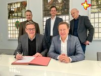 Die genannten Personen sitzen bzw.stehen im Sitzungszimmer im Rathaus und schauen in die Kamera. Die beiden Bürgermeister unterzeichnen handschriftlich einen Vertrag.