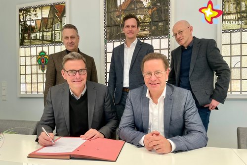 Die genannten Personen sitzen bzw.stehen im Sitzungszimmer im Rathaus und schauen in die Kamera. Die beiden Bürgermeister unterzeichnen handschriftlich einen Vertrag.