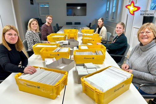 Christiane Burghardt, Kerstin Paulfeuerborn, Jan Hüttenhölscher, Maleen Henke, Emelie Maier und Astrid Karweger sitzen rund um einen großen Tisch, auf dem viele gelbe Kisten, gefüllt mit Briefen und Zetteln, stehen.