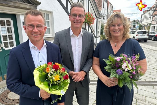 Dominik Bartels, Bürgermeister Andreas Sunder und Annette Dewenter stehen nebeneinander vor dem Rietberger Rathaus und blicken in die Kamera. Die Jubilare halten Blumensträuße in den Händen. Foto: Stadt Rietberg