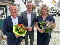 Dominik Bartels, Bürgermeister Andreas Sunder und Annette Dewenter stehen nebeneinander vor dem Rietberger Rathaus und blicken in die Kamera. Die Jubilare halten Blumensträuße in den Händen. Foto: Stadt Rietberg