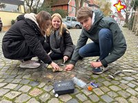 Die Schülerinnen (von links) Laila Sudermann, Felicia Scholz und Schüler Lucas Vonk hocken an der Müntestraße auf dem Boden. Mit Bürsten putzen sie die Stolpersteine, die in das Kopfsteinpflaster eingelassen sind.