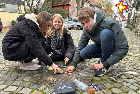 Die Schülerinnen (von links) Laila Sudermann, Felicia Scholz und Schüler Lucas Vonk hocken an der Müntestraße auf dem Boden. Mit Bürsten putzen sie die Stolpersteine, die in das Kopfsteinpflaster eingelassen sind.